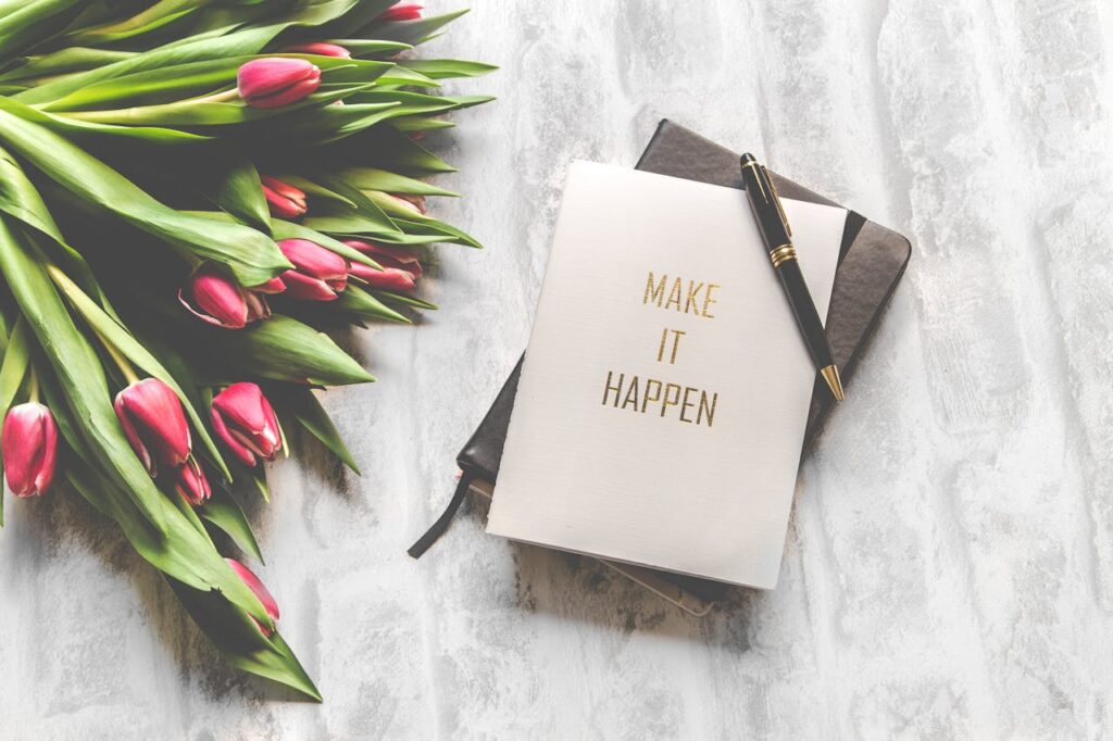 Notebook with a card reading "make it happen" and flowers.  From Pexels - https://www.pexels.com/photo/make-it-happen-book-with-black-stylus-843227/