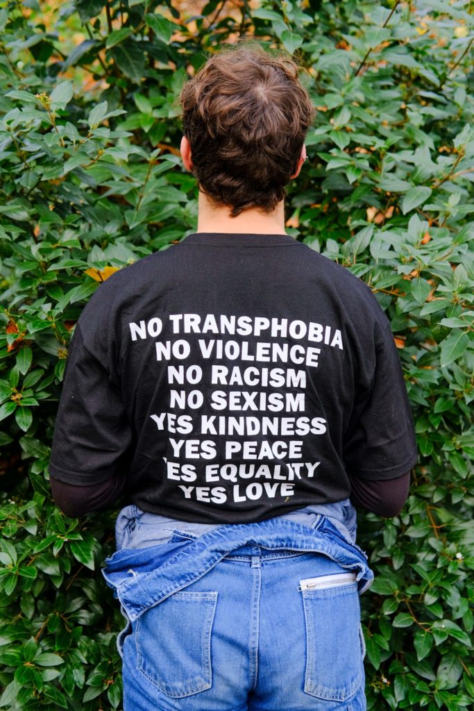 Person standing with their back to the camera wearing a shirt reading:" No transphobia, no violence, no racism, no sexism; yes kindness, yes peace, yes equality, yes love."