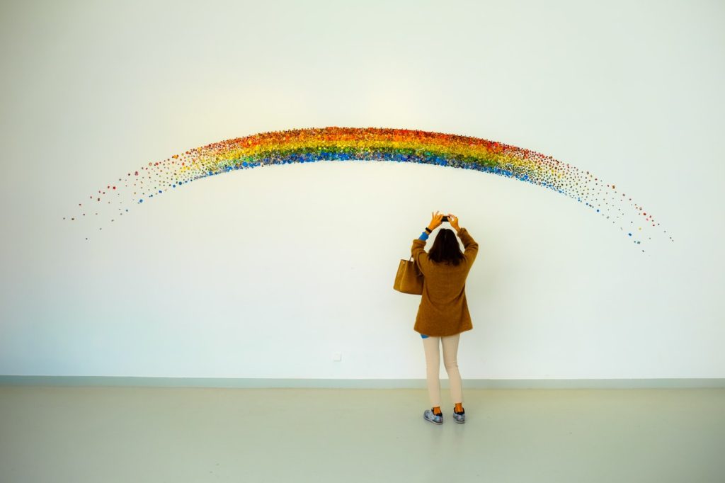 Person photographing an art installation: a rainbow comprising a curved collage of multicoloured sequins stuck onto a white wall.