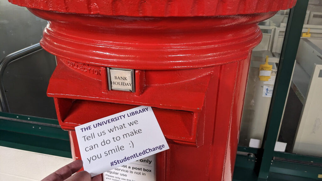Retro red cast iron postbox stood near the IT Help Desk.  A hand is just visible, in the act of posting a University Library postcard reading "Tell us what we can do to make you smile :) #StudentLedChange"